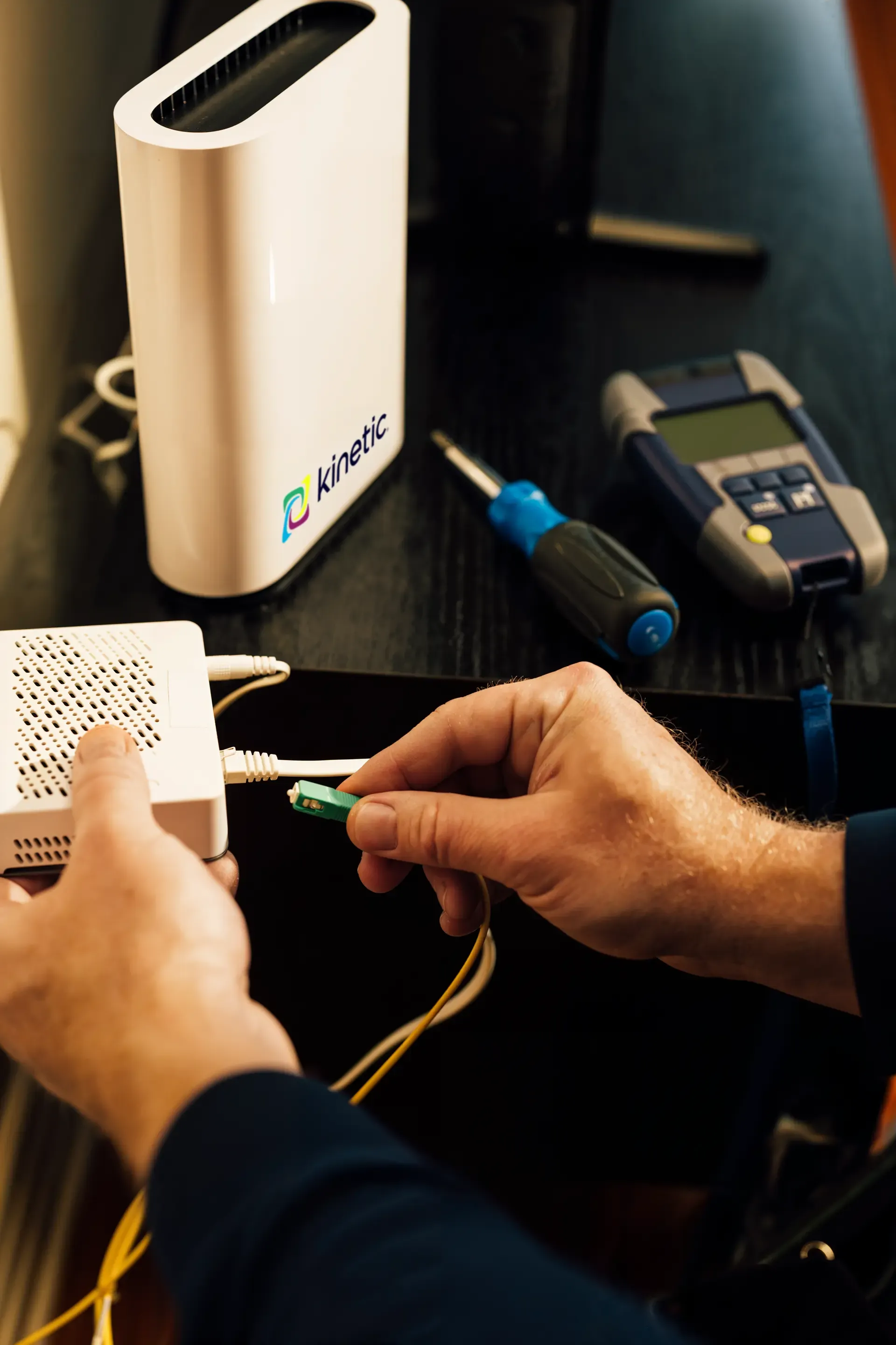 A technician's hands connecting a green fiber optic cable into a white network terminal next to a Kinetic branded router.